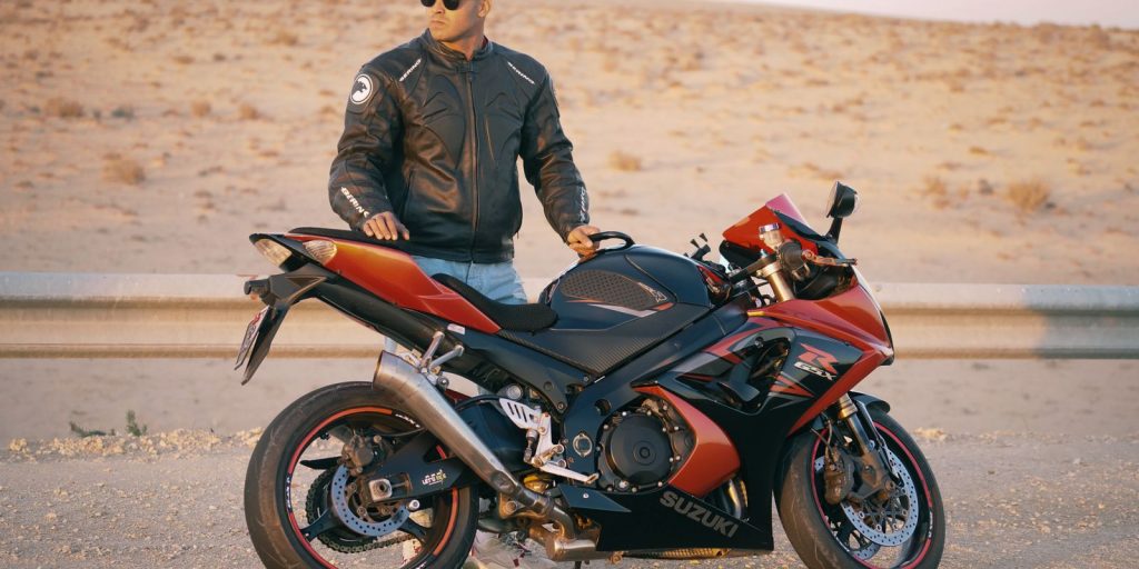 Homme en veste en cuir et casquette posant avec une moto sportive sur une route désertique