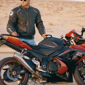 Buneri.fr | Bomber, motard ou racer : quel style de veste en cuir vous correspond ? Homme en veste en cuir et casquette posant avec une moto sportive sur une route désertique