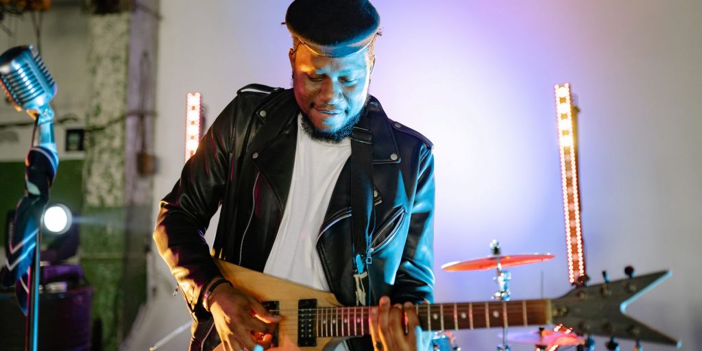 Guitariste jouant de la guitare électrique sur scène avec des lumières vives, portant une veste en cuir et une casquette plate
