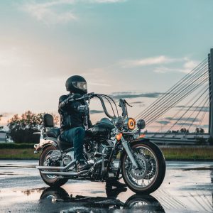 Buneri.fr | Peut-on porter une veste en cuir sous la pluie ? Motard en tenue de cuir sur une route mouillée avec un pont en arrière-plan au coucher du soleil